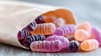 Colorful sugar coated gummy candy worms spilling from paper bag on wooden surface, selective focus on sweet jelly confectionery treats in pink, orange and purple.