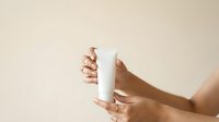 Close-up of hands holding white cylindrical product bottle against neutral background for cosmetics, skincare, or beauty packaging mockup and branding purposes.