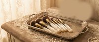 Luxury makeup brushes with wooden handles arranged on ornate vintage silver tray in elegant bedroom setting for beauty and cosmetics applications.
