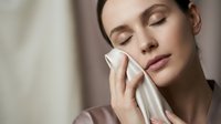 Young woman with closed eyes gently cleansing face with white cotton pad in soft natural light for skincare routine and facial care concept.