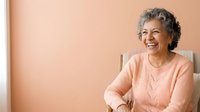 Senior woman with gray hair and warm smile wearing pink cardigan, sitting against neutral beige background. Suitable for healthcare, wellness, lifestyle and aging-related content.