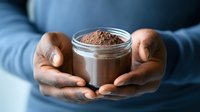 African American hands holding glass jar filled with raw cacao powder or chocolate protein supplement against blue background, healthy lifestyle concept.
