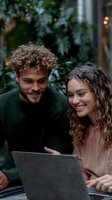 Happy young couple with curly hair laughing while using laptop computer together outdoors in cozy cafe setting with green plants in background.