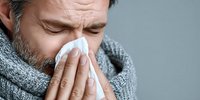 Middle-aged Caucasian man with gray scarf blowing nose into tissue, showing symptoms of cold or flu against neutral gray background.