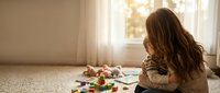 Young girl with long brown hair playing with colorful building blocks on carpet in bright living room with natural sunlight streaming through window.