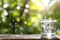 Clear water pouring into glass on wooden surface against blurred green bokeh background, splashing drops and bubbles create refreshing summer scene.