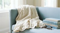 Cozy reading nook with stacked books, eyeglasses and cream throw blanket draped over modern upholstered armchair by bright window for lifestyle and home decor projects.