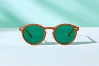 Round brown frame sunglasses with green lenses on minimal white surface against turquoise background. Modern eyewear fashion photography with soft shadows.
