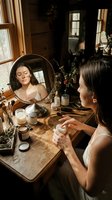 Woman with long dark hair and fair complexion applying skincare products at wooden vanity table with round mirror, cosmetics, and natural light from windows in warm-toned interior.