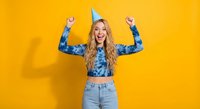 Excited young blonde woman celebrating birthday with party hat and raised arms against bright yellow background for festive occasions and celebrations.