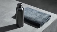 Black metal bottle with folded gray towel on concrete surface, minimalist spa and wellness product display for skincare, cosmetics, and bathroom design projects.
