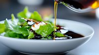 Fresh green salad leaves with balsamic vinegar dressing being poured from bottle, close up view on white ceramic plate against dark background.