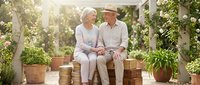 Senior couple sitting together on bench in sunny greenhouse surrounded by potted plants and climbing vines, enjoying quality time outdoors in garden setting.