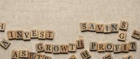 Wooden letter tiles spelling financial terms like invest, savings, growth, profit and assets scattered on textured beige background for business concepts.