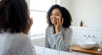 Young African American woman applying skincare cream in modern bathroom mirror reflection for daily beauty routine and self-care wellness ritual.