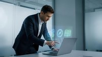 Caucasian businessman in suit working on laptop with glowing digital icons in modern office, technology and innovation concept.
