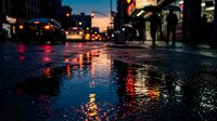 Wet urban street at dusk reflects neon lights and city traffic, creating moody atmosphere for evening scenes, nightlife backgrounds, and metropolitan settings.