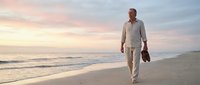 Middle-aged Caucasian man in casual beige linen clothing walking barefoot along sandy beach at sunset, carrying shoes in hand, serene coastal landscape with calm waves and golden hour lighting.