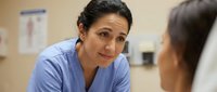 Middle-aged Hispanic female healthcare professional in blue scrubs having compassionate conversation with patient in medical office setting.