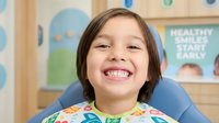 Happy young girl with brown hair smiling broadly in dental chair at pediatric dentist office, showing healthy teeth during routine checkup visit.