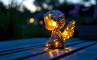 Glowing decorative angel figurine with warm LED lights inside on wooden bench at dusk, creating magical ambient atmosphere in garden setting.