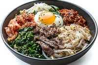Korean bibimbap bowl with marinated beef, fried egg, spinach, kimchi, rice noodles and sesame seeds served in black ceramic bowl on white background.