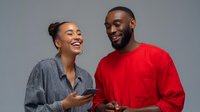 Two young adults, a woman of mixed ethnicity with hair in a bun wearing a gray shirt and a man of African descent wearing a red shirt, smile while holding smartphones and looking at each other against a neutral gray background. Perfect for communication, technology, and lifestyle content.