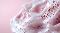 Creamy pink foam with delicate bubbles and soft swirls creates dreamy abstract texture against rosy background, macro photography capturing liquid movement.