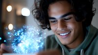Young Hispanic man with curly hair smiling while looking at glowing blue magical light effect emerging from smartphone in dark room, creating ethereal atmosphere.