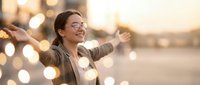 Young Caucasian businesswoman with glasses smiling joyfully, arms outstretched outdoors at sunset with golden bokeh lights background.