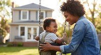 African American mother pushing happy toddler son on swing in backyard during golden hour sunset creating joyful family bonding moment outdoors at home.