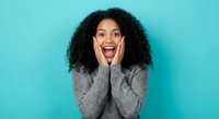 Excited young African American woman with curly hair expressing surprise and joy, hands on cheeks, wearing gray sweater on turquoise background.