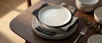 Elegant table setting with stacked white and black plates, linen napkin, and cutlery on wooden surface, suitable for dining, hospitality, and restaurant design projects.