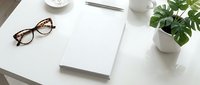 Clean minimalist workspace with eyeglasses, notebook, pen and green plant on white desk for productivity and organization.