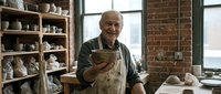 Senior male artisan of Caucasian ethnicity wearing apron, holding clay vessel in pottery studio with shelves of ceramics and brick walls, demonstrating traditional craftsmanship and creative hobby.