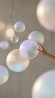 Hand touching a glowing iridescent sphere among floating pearlescent bubbles against a concrete wall, dreamy pastel tones.