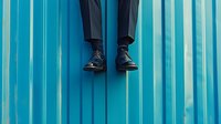 Minimalist composition of black dress shoes and suit pants against turquoise corrugated metal wall, legs dangling in surreal floating position for creative design.