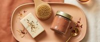 Flat lay of spa skincare essentials on a pink tray, featuring a dry brush, herbal soap bar, clay mask jar, and golden spoon with rose petals.