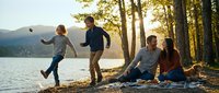 Young family enjoying outdoor recreation by a mountain lake at sunset, with children playing and parents relaxing on the shore among trees.