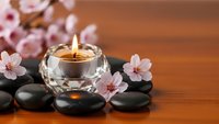Zen spa composition with burning candle in crystal holder, black stones and delicate pink cherry blossoms on warm wooden surface for relaxation.