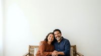 Mature couple sitting together on wooden bench against white wall, smiling at camera. Hispanic man and woman in casual clothing posing for portrait.