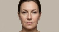 Professional portrait of confident middle-aged Caucasian businesswoman with natural makeup and pulled-back hair against neutral beige background.