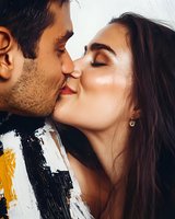 Young couple kissing in close-up, man with dark hair and facial stubble wearing striped shirt, woman with long dark hair and earring, romantic moment with natural lighting.