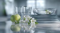 Fresh green apple and delicate white spring blossom alongside stacked clear glass plates and drinking glasses on reflective surface with soft bokeh background.