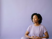 Young woman of African descent with shoulder-length curly black hair, wearing a lavender t-shirt, sitting against a purple background with hands resting on lap, expressing calm contentment and relaxation for wellness and lifestyle concepts.