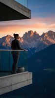 Woman standing on modern concrete balcony overlooking dramatic mountain peaks at sunset, contemplating scenic alpine landscape with golden sky and distant valleys below.