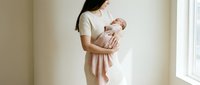 Young Asian mother in white dress gently holds sleeping newborn wrapped in pink blanket near bright window, soft natural light.