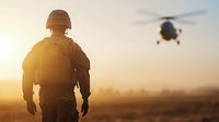 Silhouette of military soldier in combat gear watching helicopter at sunset, dramatic orange sky and desert landscape creates powerful military scene.