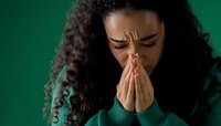 Young African American woman with curly hair praying or showing distress against green background, expressing deep emotion and vulnerability in contemplative pose.