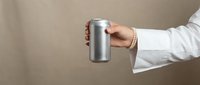 Professional woman hand holding silver aluminum beverage can against neutral beige background for product presentation and marketing materials.